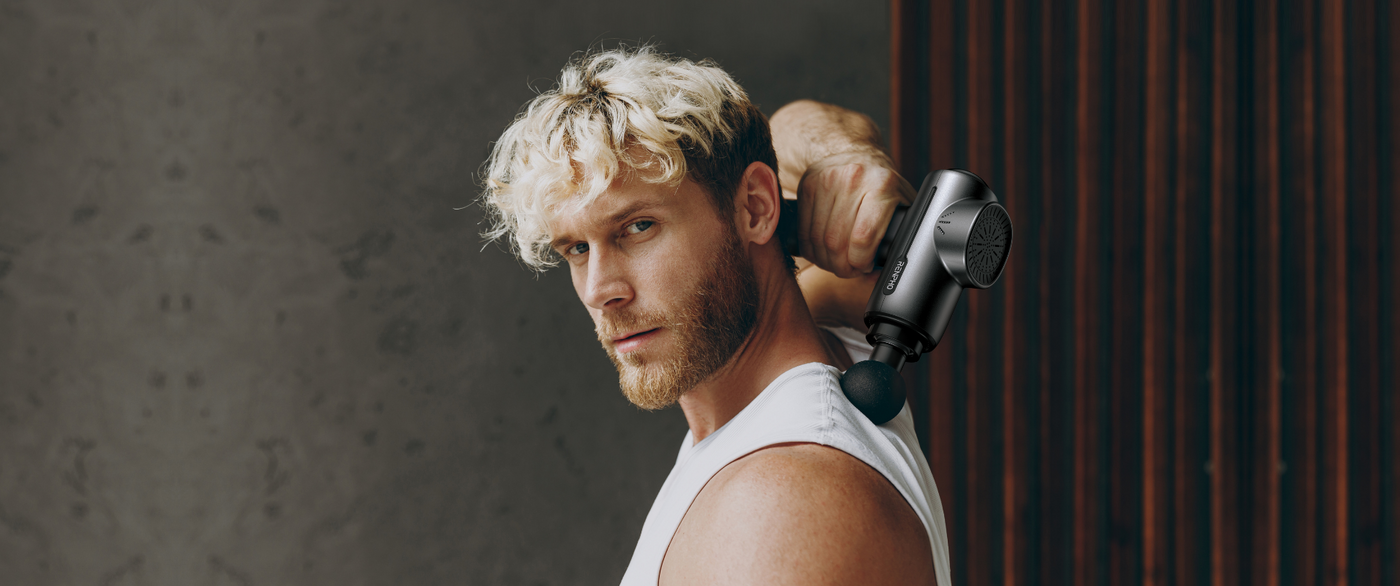 A muscular man with curly blonde hair and a beard uses a black massage gun on his shoulder. He wears a sleeveless white shirt and looks intently at the camera. The background features a textured concrete wall and vertical wooden slats.