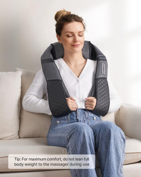 A woman sitting on a couch, relaxing with a U-Neck 2 Neck & Shoulders Massager. She is wearing a white sweater and jeans, and has her eyes closed, appearing content.